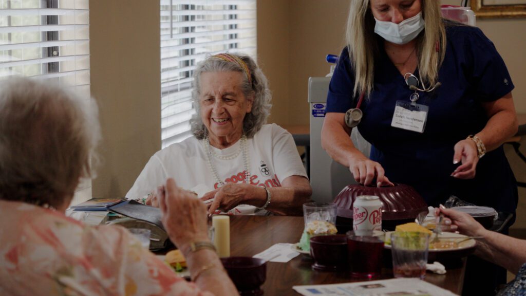 A woman smiles as a nurse assists her with dinner at a skilled nursing facility